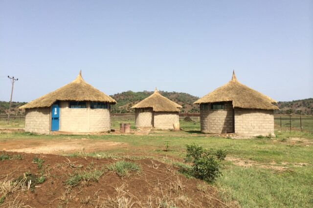 A circular stone building with a thatched roof, with similar buildings on either side of it.