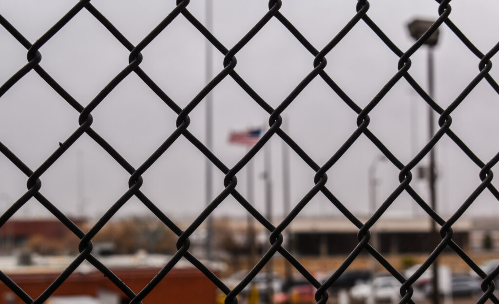 A wire fence with out of focus buildings behind it.