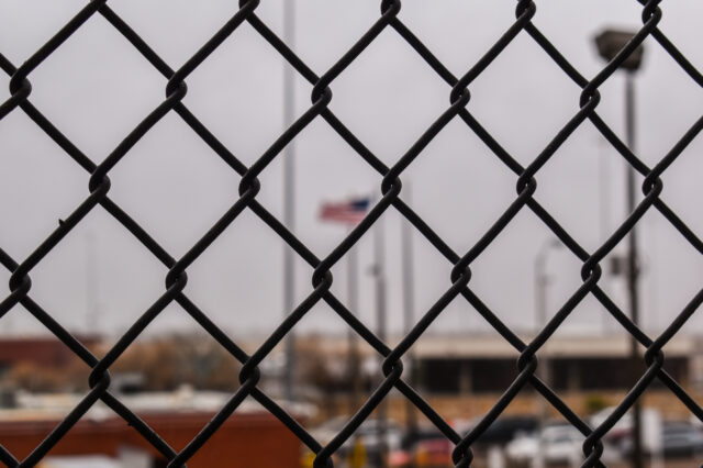 A wire fence with out of focus buildings behind it.