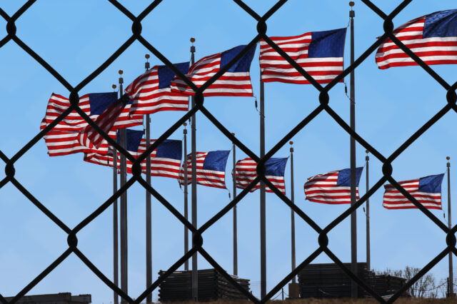 A wire fence with several American flags waving behind it.