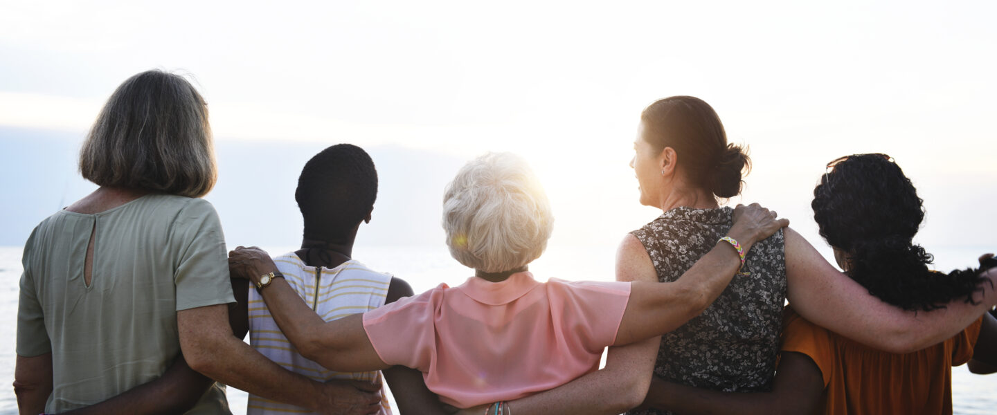 A group of five women do a linear group hug, facing away from the camera.