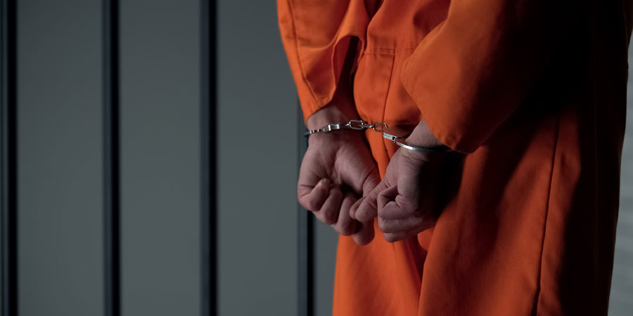 A man in an orange jumpsuit and handcuffs stands in front of prison bars with hands behind his back.