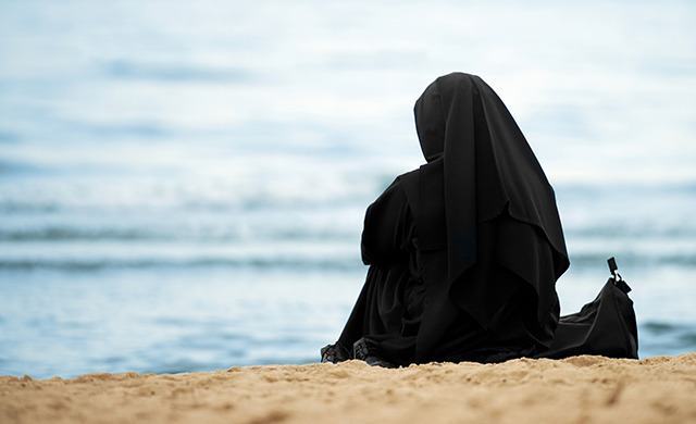 A woman wearing all black with her hair covered, sits on a sandy beach looking at the ocean. Her back faces the camera.