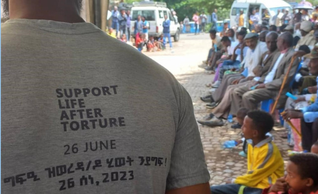 The back of a shirt reads Support Life After Torture, June 26, and to the left a group of community members are seen sitting outside